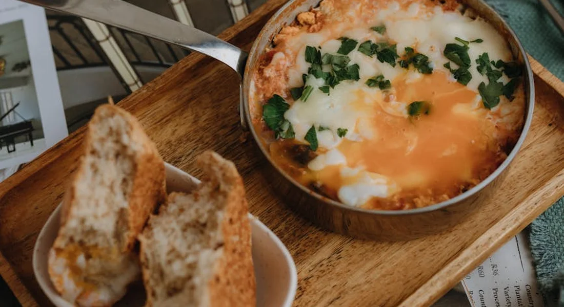 A cast iron skillet with sunny-side-up eggs and chopped herbs sits on a wooden board, with slices of crusty bread nearby.