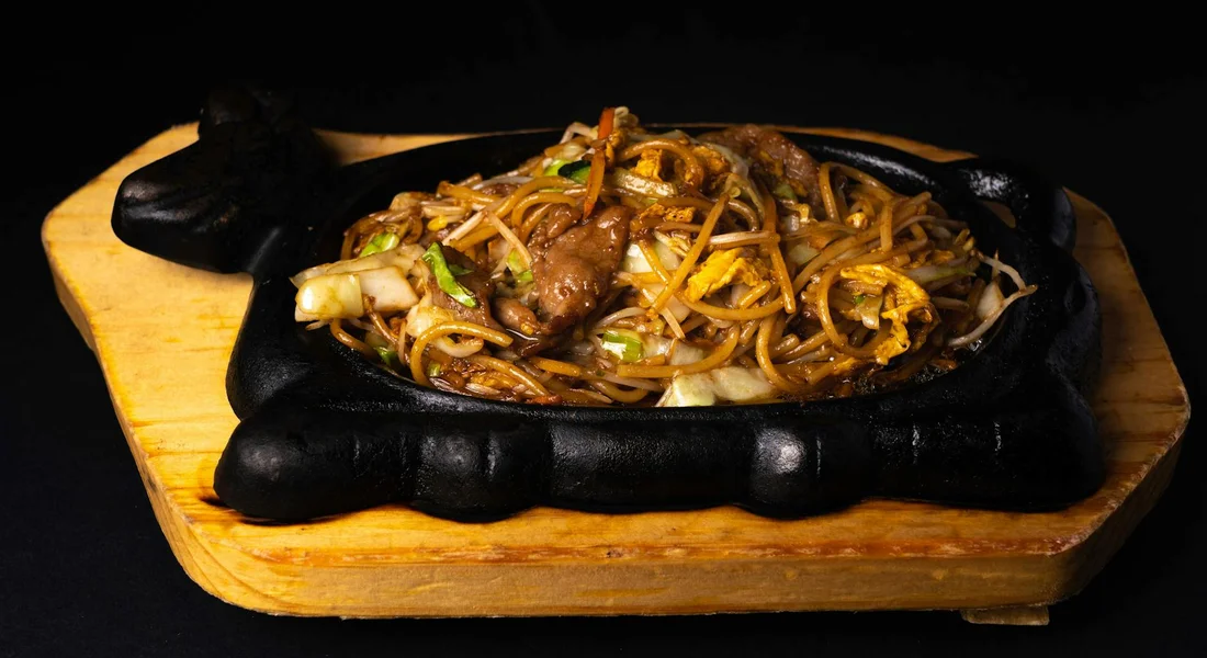 Stir-fried noodles with meat and vegetables on a black cast-iron skillet placed on a wooden board.