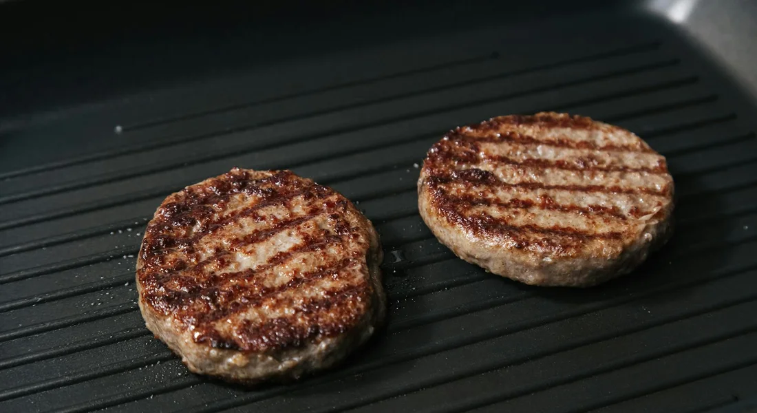 Two beef burger patties with grill marks cooking on a ridged cast-iron grill pan.