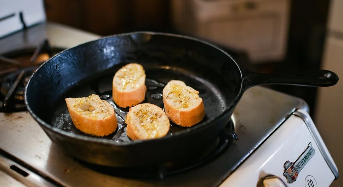 Seasoned cast iron skillet on a kitchen stove with four slices of bread in the pan, highlighting where to inspect for maker marks and brand identifiers.