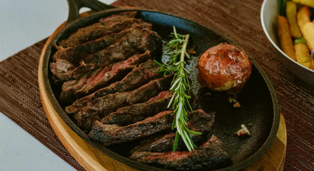 Sliced steak arranged in a cast iron skillet with a sprig of rosemary, a roasted onion, and a side of fries in a white bowl nearby.