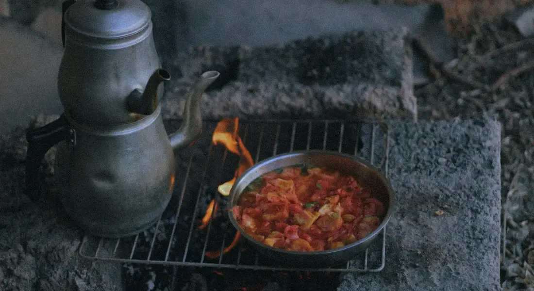Close-up of a vintage kettle and a small metal skillet on a grill over glowing coals outdoors.