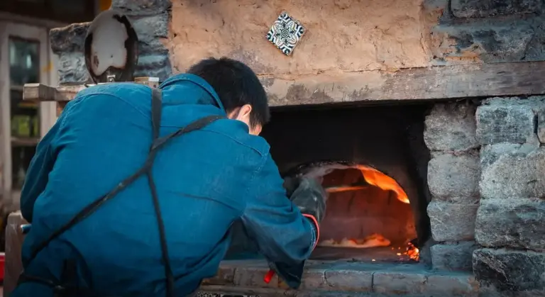 Person in a blue jacket leaning into a brick wood-fired oven with orange flames visible inside